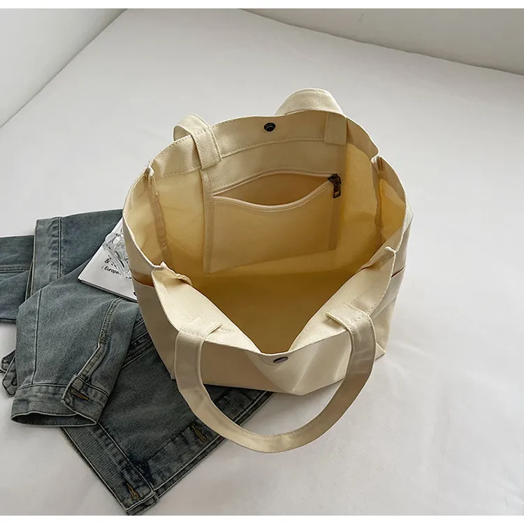 Open cream canvas tote showing its internal zip pocket, snap closure and stitched handle attachments